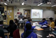 A classroom full with students and a teacher. Teacher is teaching the students with the presentation in front of the classroom.