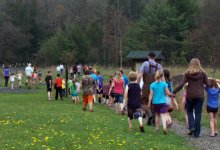 Group of teacehrs and students are on a field trip. They're walking in a green field.