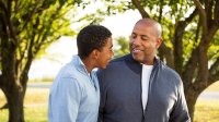 Standing outside between two trees, a father and son are looking at each other, smiling, and hugging each other with one arm. 