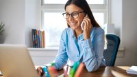 A young female teacher wearing glasses is at her desk smiling and on the phone.