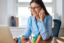 A young female teacher wearing glasses is at her desk smiling and on the phone.