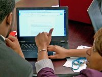 Teacher crouching down at a desk helping a student with his composition on a computer