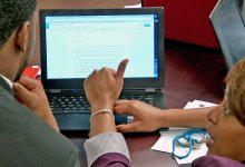 Teacher crouching down at a desk helping a student with his composition on a computer