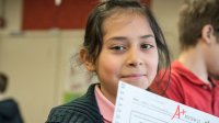 A young girl is standing in a classroom, holding up a piece of paper that shows an A plus drawn in red marker. 