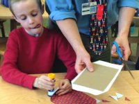 Boy sitting with tongue out gluing with great concentration; teacher standing next to him using scissors