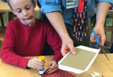 Boy sitting with tongue out gluing with great concentration; teacher standing next to him using scissors