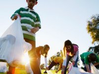 A group of young teens are outside in the sun picking up garbage from the grass and putting it into white trash bags.