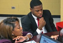 A teenage boy is sitting a table looking at a desktop computer screen. A female teacher is kneeling on the floor beside him, also looking at the computer, and talking. 