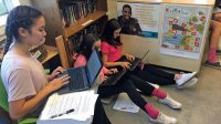 Girls work in class while sitting on chairs or the floor.