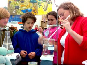 Teacher stirring a liquid in a beaker on a Bunsen burner with three students looking on