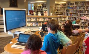 A photo of a group of students gathered around a computer workstation, each at their own laptop.