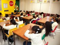 A group of young students are sitting in a classroom looking towards an adult male. At the front of the class, the wall is yellow. A red and white stop sign is hanging on the wall next to another sign that says, "yield to pedestrian in crosswalk."