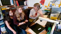 two high school aged students working with a teacher around a computer