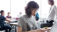 A female high school student works at a computer as other students talk to the teacher in the background.