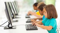 A row of kids work at computers in their class.