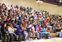 In the multipurpose room, a large group of middle school students fill one side of the bleachers, some clapping.