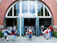 Eight elementary-age students are smiling, running out of the school building through three opened doors. 