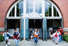 Eight elementary-age students are smiling, running out of the school building through three opened doors. 