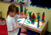 A young girl with long hair in a white t-shirt and blue shorts is sitting at a kid-sized table with colorful, clear, Lego-like building pieces. On the table are variations of different towers.