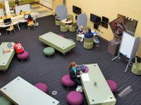 Teachers and students are sitting in a uniquely designed and furnished classroom with different sitting chairs and different sized and shaped tables.