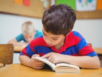 A young boy in a large-striped red and blue polar shirt is sitting at his desk with his head down in a book.