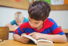 A young boy in a large-striped red and blue polar shirt is sitting at his desk with his head down in a book.