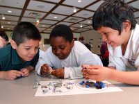 Three young boys are hovering over their classroom table, smiling. They're holding and looking at nuts, bolts, and screws that are lying on a piece of lamented paper with two circles drawn on it.