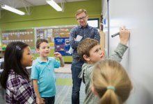 Elementary school students working together at a whiteboard with their teacher observing