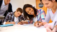 Three teenage students work together, writing and smiling, while their teacher assists. 