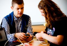 Two teenagers are sitting at a table with a laptop, one is holding a cell phone charger and the other is holding a cell phone, connecting it to a white wire.