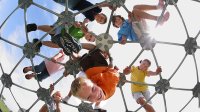 Eight young students are outside on top of a metal, geodesic dome at a playground, looking down. 