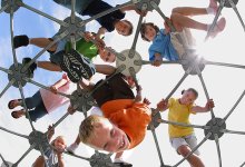 Eight young students are outside on top of a metal, geodesic dome at a playground, looking down. 