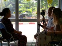 Three adults are sitting across from an old woman in a classroom, filming her. One of them is holding a camera on a tripod, and another is holding a long microphone.