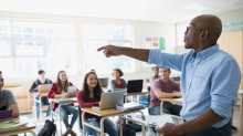 Photo of a male teacher calling on a student in a classroom filled with smiling, seated high schoolers