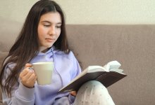 Teen girl reads a book while sitting on a couch and drinking from a mug