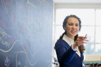 Teacher standing next to a blackboard that has a diagram drawn on it. She is pointing off-camera.