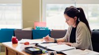 Teacher sitting at desk in classroom writing. 