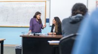 High school student speaking with teacher at teacher;s desk in classroom. 