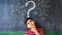 A young boy stands thinking in front of a blackboard covered with question marks.