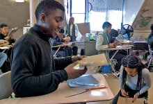 A high school boy is sitting at his desk, smiling, folding a piece of cardboard with pieces of tape on it. The class is filled with students at their desks or on the floor cutting and taping together pieces of cardboard.