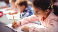 A young girl in the foreground and other kids beyond her write in notebooks.