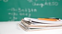 A stack of four notebooks are on a classroom desk with three pencils lying on top of them. A blurred out chalkboard is shown in the background.