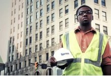 Boy wearing a yellow safety vest holding his hard hat, standing on a busy street in front of a very large building