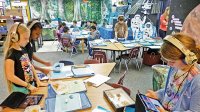 Tablets and lamented information sheets next to boxes filled with marine life span across nine classroom tables. Seven students are sitting at one table, two students are standing together, and two others wearing headphones are working individually.