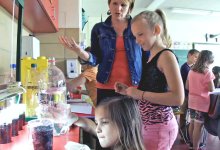 Teacher standing talking with a student as they watch a girl pouring liquid into cups at a counter