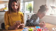 A mom and daughter are sitting beside each other at the kitchen table. The mom is looking at her phone while the daughter is playing with beads.