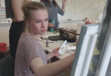 A female teen is sitting in an art class, holding a long paint brush, painting on a canvas.