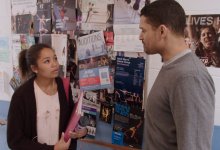 A female teen is standing in a school hallway talking to a male teacher; there are dance fliers on a bulletin board on the wall beside them.