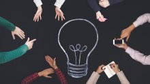 Top view of six pairs of hands around a chalk drawing of a lightbulb.