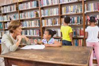 A librarian talking to a young girl at a table while two students browse the stacks behind them